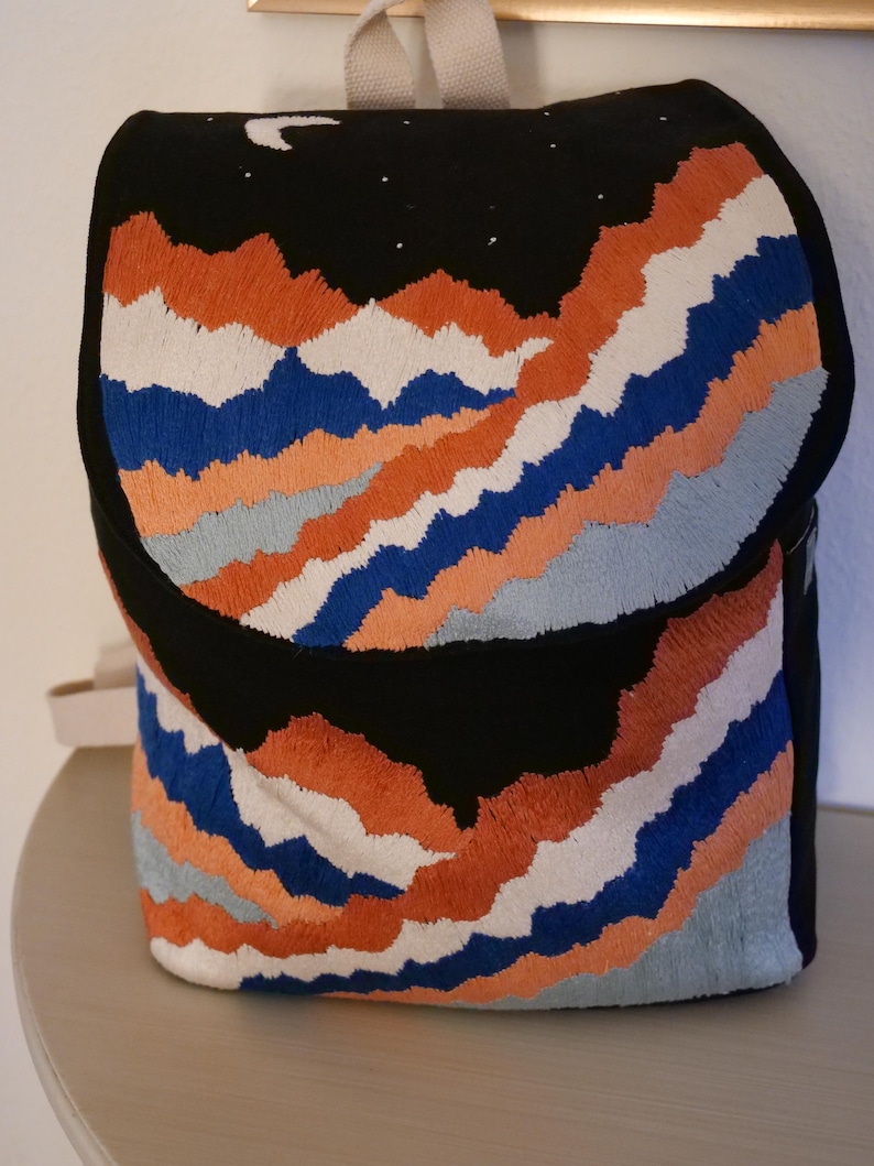 May include: Black backpack with embroidered mountain range design in orange, blue, white, and gray. The top flap features a crescent moon and stars. The backpack has a beige handle and straps.