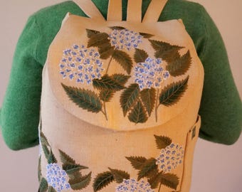 May include: A beige backpack with a flap closure, adorned with a floral design featuring blue hydrangea blooms and green leaves. The backpack has adjustable straps and a side pocket, offering a blend of style and functionality.