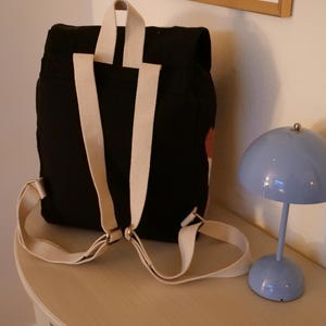 May include: Black backpack with cream-colored straps and a small red and white design. The backpack is on a light-colored surface next to a small, blue, dome-shaped lamp.