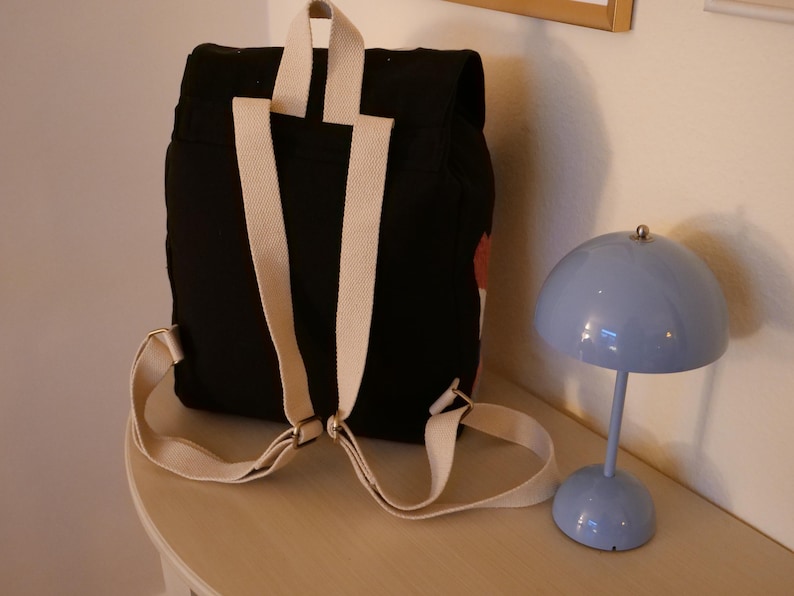 May include: Black backpack with cream-colored straps and a small red and white design. The backpack is on a light-colored surface next to a small, blue, dome-shaped lamp.