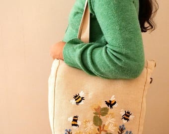 May include: A beige canvas tote bag with embroidered bees and flowers. The bag has a long strap and is being held by a person wearing a green sweater.