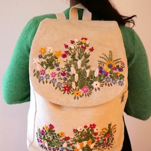 May include: A beige canvas backpack with a flap closure. The backpack is embroidered with colourful flowers and greenery.