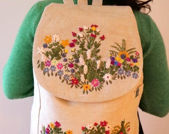 May include: A beige canvas backpack with a flap closure. The backpack is embroidered with colorful flowers and greenery.