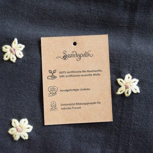 May include: A close-up shot of a dark blue fabric with three embroidered flowers and a brown tag. The tag reads "Sundapath" and lists details about the product, including GOTS certified organic cotton and handmade items.