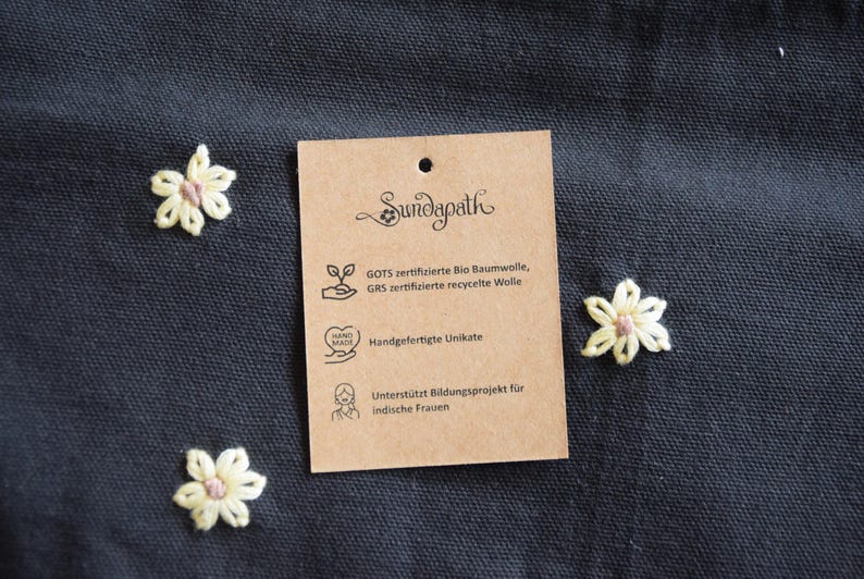 May include: A close-up shot of a dark blue fabric with three embroidered flowers and a brown tag. The tag reads "Sundapath" and lists details about the product, including GOTS certified organic cotton and handmade items.