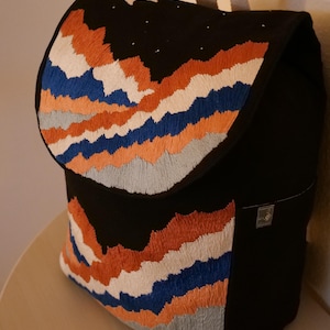 May include: Black backpack with an embroidered mountain range design in orange, white, blue, and gray. The flap features a night sky with a crescent moon and stars. The bag has a top handle and a small tag.