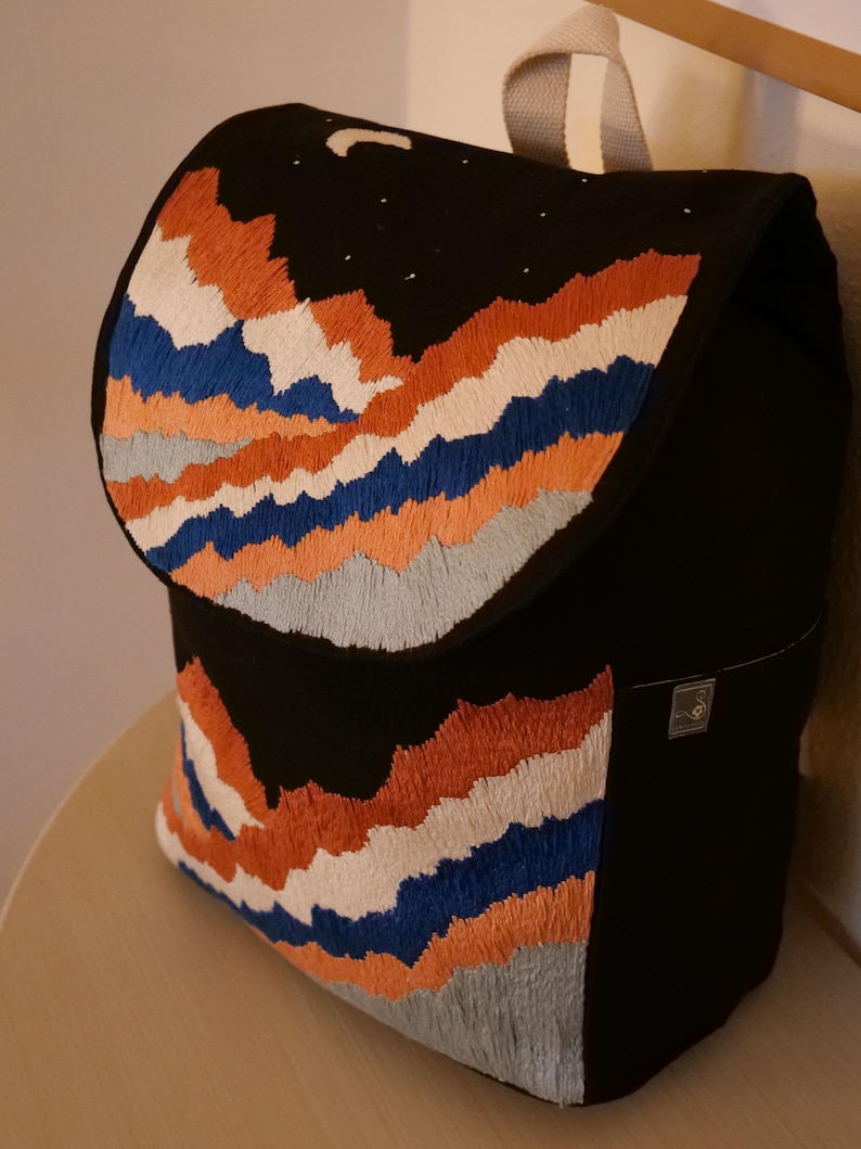 May include: Black backpack with an embroidered mountain range design in orange, white, blue, and gray. The flap features a night sky with a crescent moon and stars. The bag has a top handle and a small tag.