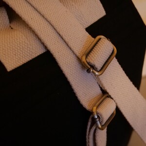 May include: Close-up of a black canvas bag with off-white canvas straps and antique brass-colored metal buckles. The straps are adjustable and attached to the bag.