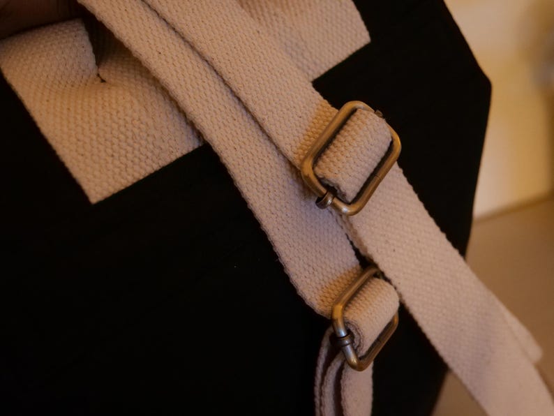 May include: Close-up of a black canvas bag with off-white canvas straps and antique brass-colored metal buckles. The straps are adjustable and attached to the bag.