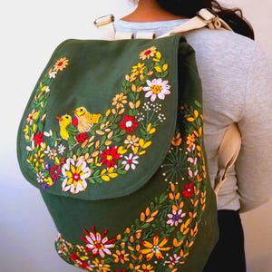 May include: A green canvas backpack with embroidered flowers and birds in shades of yellow, red, pink, and white.