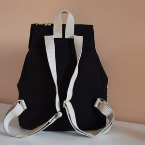 Pode incluir: Mochila de lona preta com al&ccedil;as brancas. A mochila tem fecho de aba e um bolso com z&iacute;per na frente.