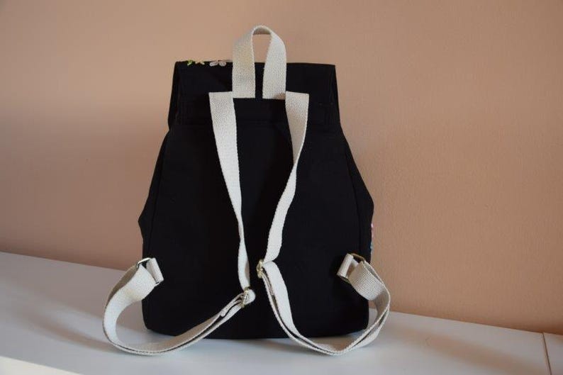 Pode incluir: Mochila de lona preta com al&ccedil;as brancas. A mochila tem fecho de aba e um bolso com z&iacute;per na frente.