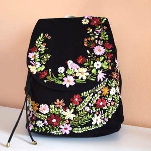 Pode incluir: Mochila preta com um design de bordado floral. O bordado apresenta flores rosa, brancas, amarelas e vermelhas, bem como folhas verdes e um pequeno p&aacute;ssaro.