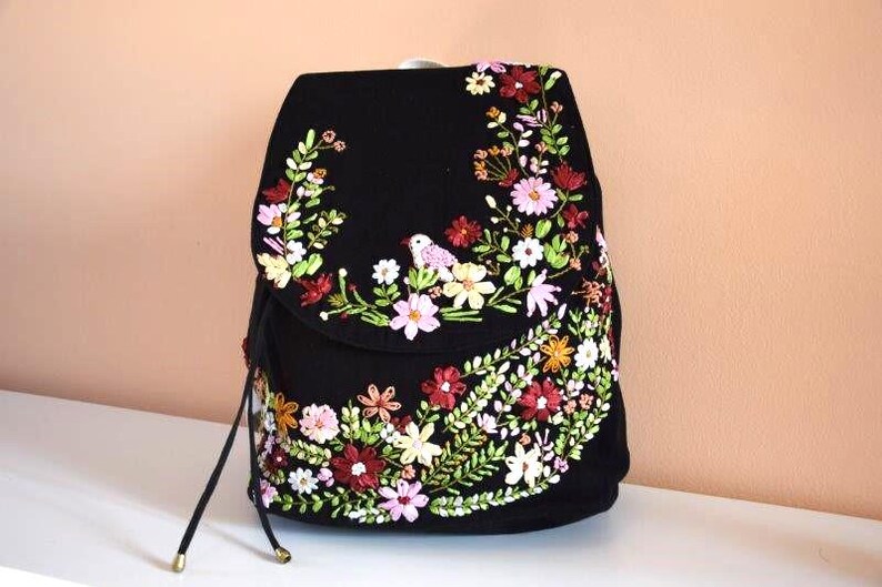 Pode incluir: Mochila preta com um design de bordado floral. O bordado apresenta flores rosa, brancas, amarelas e vermelhas, bem como folhas verdes e um pequeno p&aacute;ssaro.
