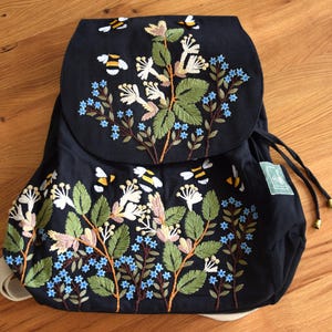 Puede incluir: Mochila negra con diseño bordado de flores y abejas. El bordado presenta detalles en blanco, azul, verde y amarillo. La mochila tiene cierre de cordón y correas ajustables. La bolsa es de tela oscura.