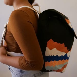 May include: Black backpack with a colorful mountain range design. The backpack features a flap closure and white straps. The mountain design includes orange, blue, and white accents. The straps are white.