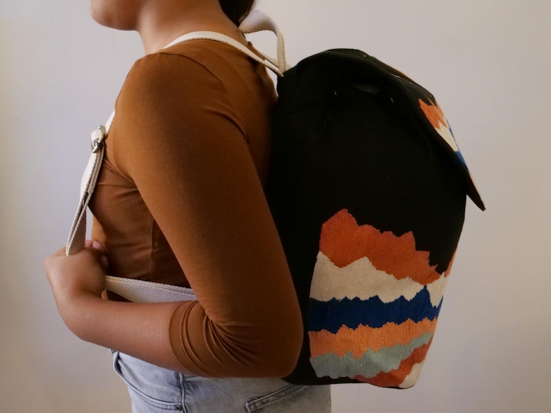 May include: Black backpack with a colorful mountain range design. The backpack features a flap closure and white straps. The mountain design includes orange, blue, and white accents. The straps are white.