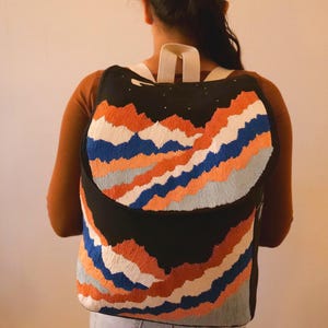 Könnte beinhalten: Schwarzer Rucksack mit einem gestickten Bergketten-Design in Orange, Blau und Weiß. Der Rucksack hat weiße Träger und eine schwarze Oberseite mit kleinen weißen Sterndetails. Die Tasche wird von einer Person getragen.