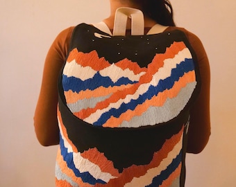 May include: Black backpack with an embroidered mountain range design in orange, blue, and white. The backpack has white straps and a black top with small white star details. The bag is worn by a person.