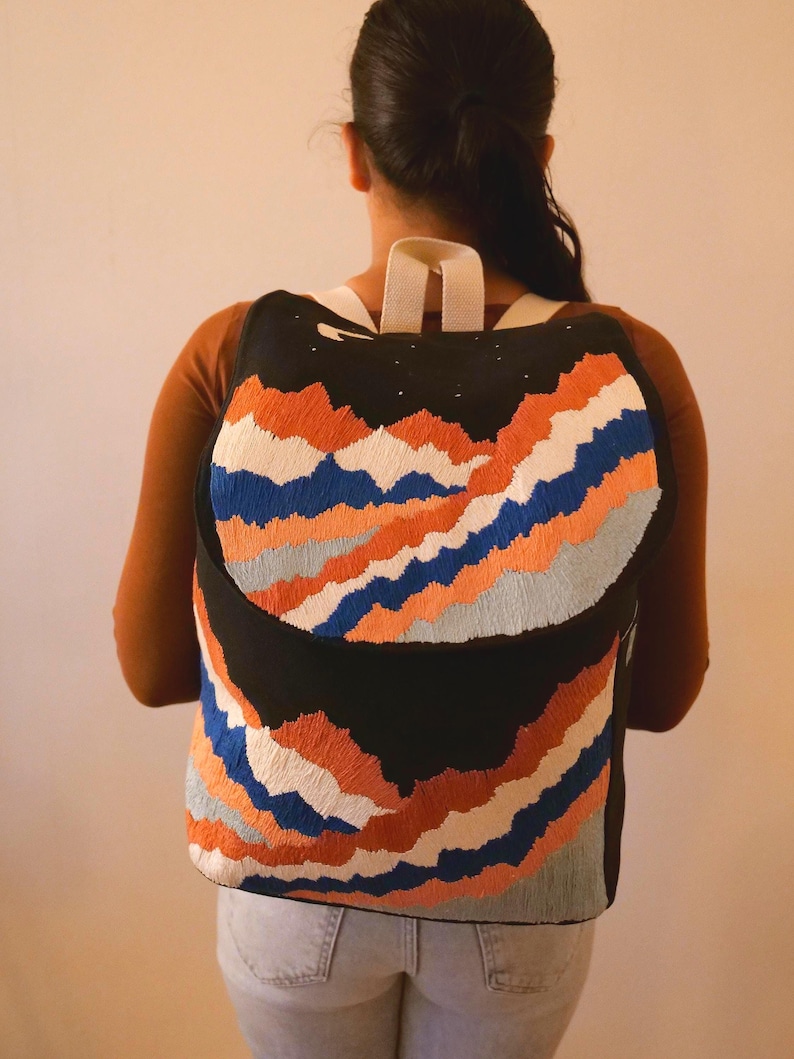May include: Black backpack with an embroidered mountain range design in orange, blue, and white. The backpack has white straps and a black top with small white star details. The bag is worn by a person.