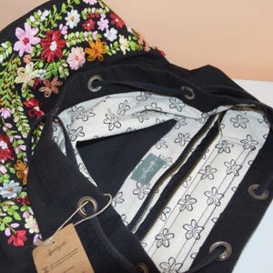 Pode incluir: Sacola tote de lona preta com um design de bordado floral. A bolsa tem fecho de correr e um topo com cord&atilde;o.