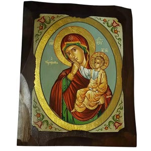 Può includere: Icona religiosa raffigurante la Vergine Maria con il Bambino Gesù, incorniciata in legno scuro. L'icona ha un bordo dorato e le figure sono vestite di rosso, verde e oro. Lo sfondo è azzurro.