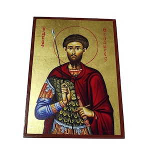 Pode incluir: Um ícone religioso com um homem com uma auréola, segurando uma lança e uma espada. O homem está vestido com roupas vermelhas e azuis, sobre um fundo dourado. O ícone tem texto grego nas laterais e é emoldurado por uma borda marrom escura.