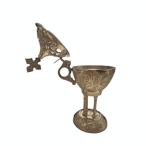 May include: A silver-colored censer with an open lid and a cross-shaped handle. The censer has intricate floral patterns and stands on a small, oval base. The lid is partially open, revealing the inside of the censer.
