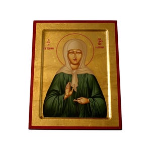 May include: A religious icon featuring a woman in a green robe and white head covering, with a gold background and red frame. The woman has a halo and is depicted with a serene expression. The icon has text in an unknown language.