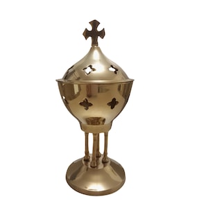 May include: A gold-colored censer with a cross-shaped finial. The censer has a round base, three legs, and a domed lid with decorative cutouts. The metal has a polished finish, reflecting light.
