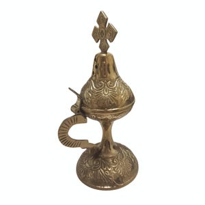 May include: A gold-colored metal incense burner with intricate floral and geometric patterns. The burner features a handle, a hinged lid, and a cross-shaped finial. The base is round and decorated with similar patterns.