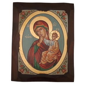 Può includere: Icona religiosa raffigurante la Vergine Maria con il Bambino Gesù, incorniciata da un'aureola dorata e da una cornice in legno scuro. L'opera d'arte ha un aspetto vintage e invecchiato, con colori sbiaditi e usura visibile.