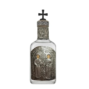 May include: A clear glass bottle featuring a silver-toned metal plaque on the front depicting two figures in robes with halos. The bottle neck is adorned with ornate silver metalwork and topped with a dark, cross-shaped stopper.