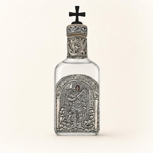 May include: A clear glass bottle featuring a silver-toned metal plaque depicting an angel with wings and a sword. The bottle has a decorative silver neck band with scrollwork and a black cross-shaped stopper at the top of the bottle.