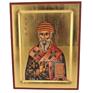 Könnte beinhalten: Eine rechteckige religiöse Ikone mit einem bärtigen Heiligen, der eine gemusterte Kopfbedeckung und ein rotes Gewand mit schwarzen Kreuzen trägt. Die Figur hält ein Buch und segnet mit der Hand vor einem goldenen Hintergrund.