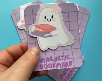 May include: A magnetic bookmark featuring a white ghost character with a small pink bow, holding a stack of two books. The bookmark is attached to a light purple rectangular card with a dark purple grid pattern and the text MAGNETIC BOOKMARK.