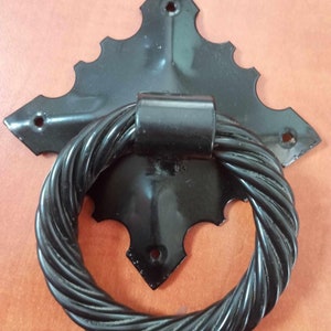 May include: Black metal door knocker with a twisted rope design. The knocker is attached to a black metal plate with a decorative design.