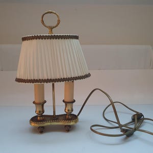 Puede incluir: Una lámpara de mesa de latón con una pantalla de lámpara plisada blanca y un ribete marrón. La lámpara tiene dos bombillas de estilo vela y un cable con un enchufe.