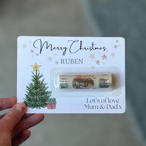 May include: A white Christmas card with the text "Merry Christmas" and "Ruben". The card features a watercolor Christmas tree, gifts, and stars. A small Christmas cracker is visible in a clear plastic case.
