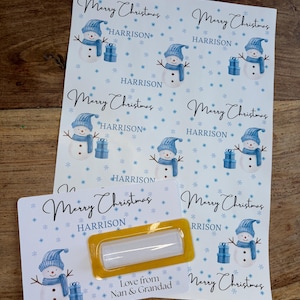 May include: Sheets of Christmas stickers with a snowman design. Each sticker features a snowman wearing a blue hat and scarf, holding a gift. The stickers are personalized with the name "HARRISON" and the text "Merry Christmas".