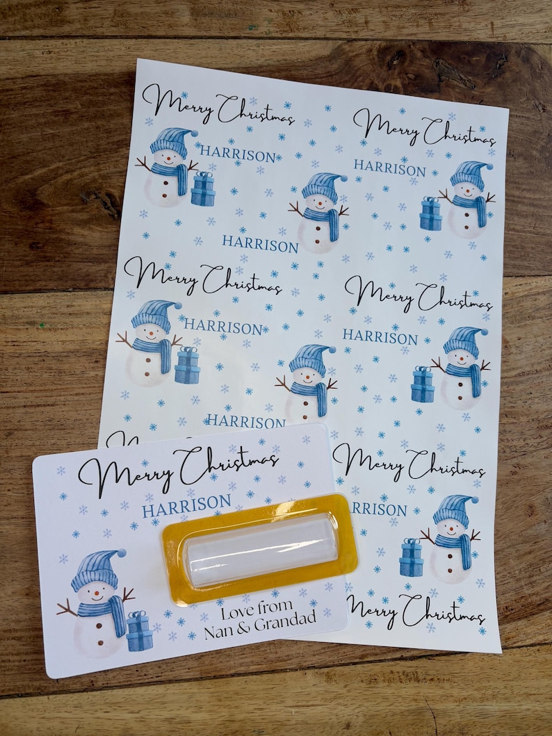 May include: Sheets of Christmas stickers with a snowman design. Each sticker features a snowman wearing a blue hat and scarf, holding a gift. The stickers are personalized with the name "HARRISON" and the text "Merry Christmas".