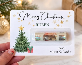 Puede incluir: Una tarjeta de regalo navideña rectangular blanca con el texto Merry Christmas y RUBEN en negro y gris. Se ilustra un árbol de Navidad verde con una estrella dorada y un regalo rojo. Un pequeño objeto cilíndrico está en plástico.