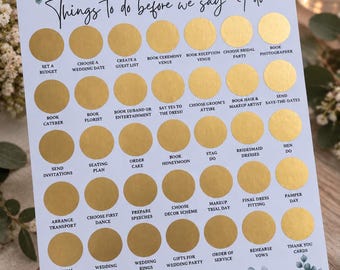 May include: A white wedding planning checklist titled Things To Do before we say I do, featuring gold foil scratch-off circles over task labels. The top and bottom borders are decorated with illustrations of green eucalyptus leaves and white roses.