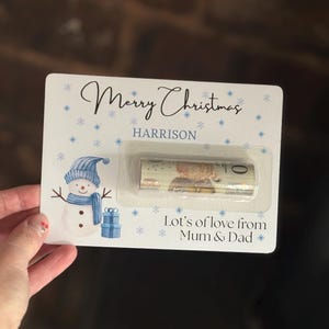 May include: A Christmas gift card with a snowman illustration, snowflakes, and the text "Merry Christmas HARRISON". The card includes a rolled-up bill in a clear plastic holder, with the message "Lot's of love from Mum & Dad".