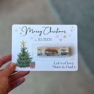 May include: A Christmas card with the words "Merry Christmas" and "Ruben" printed on it. The card features a Christmas tree illustration, star accents, and the message "Let's of love Mum & Dad x". A small gift is included.
