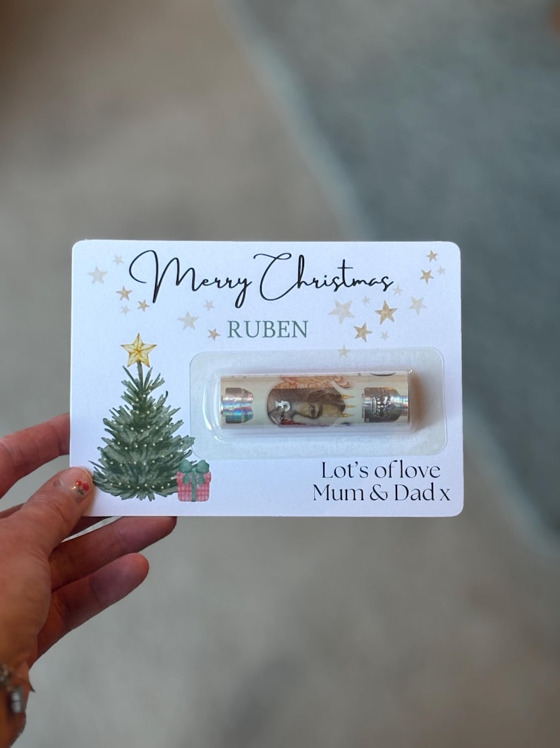 May include: A Christmas card with the words "Merry Christmas" and "Ruben" printed on it. The card features a Christmas tree illustration, star accents, and the message "Let's of love Mum & Dad x". A small gift is included.