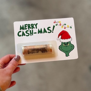Puede incluir: Una tarjeta blanca con el texto "MERRY CASH-MAS!" en verde, con un dibujo animado del Grinch con un gorro de Pap&aacute; Noel. Un billete enrollado es visible en una funda de pl&aacute;stico transparente. Tarjeta de regalo con tem&aacute;tica navide&ntilde;a.