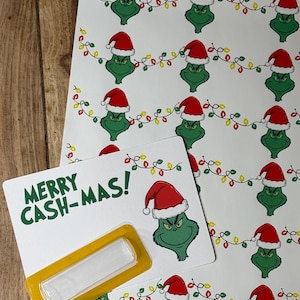 Puede incluir: Hojas de papel blanco con un patr&oacute;n repetido de un personaje de dibujos animados verde con un gorro de Pap&aacute; Noel y luces de cadena. Una hoja tiene el texto "MERRY CASH-MAS!" en verde, con un rect&aacute;ngulo amarillo.