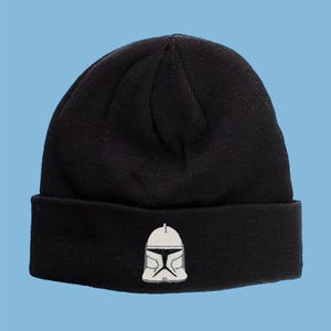 Puede incluir: Gorro de punto negro con un diseño de casco bordado en blanco y gris en la parte delantera. El gorro tiene un dobladillo doblado y está sobre un fondo azul claro. Un gorro cálido para el frío.