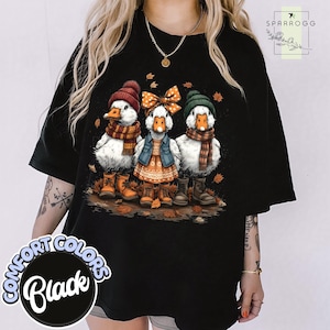 May include: Black Comfort Colors t-shirt with a charming illustration of three ducks in autumn outfits. The ducks are wearing hats, scarves, and boots, amidst fallen leaves. The shirt has the text "Comfort Colors Black".